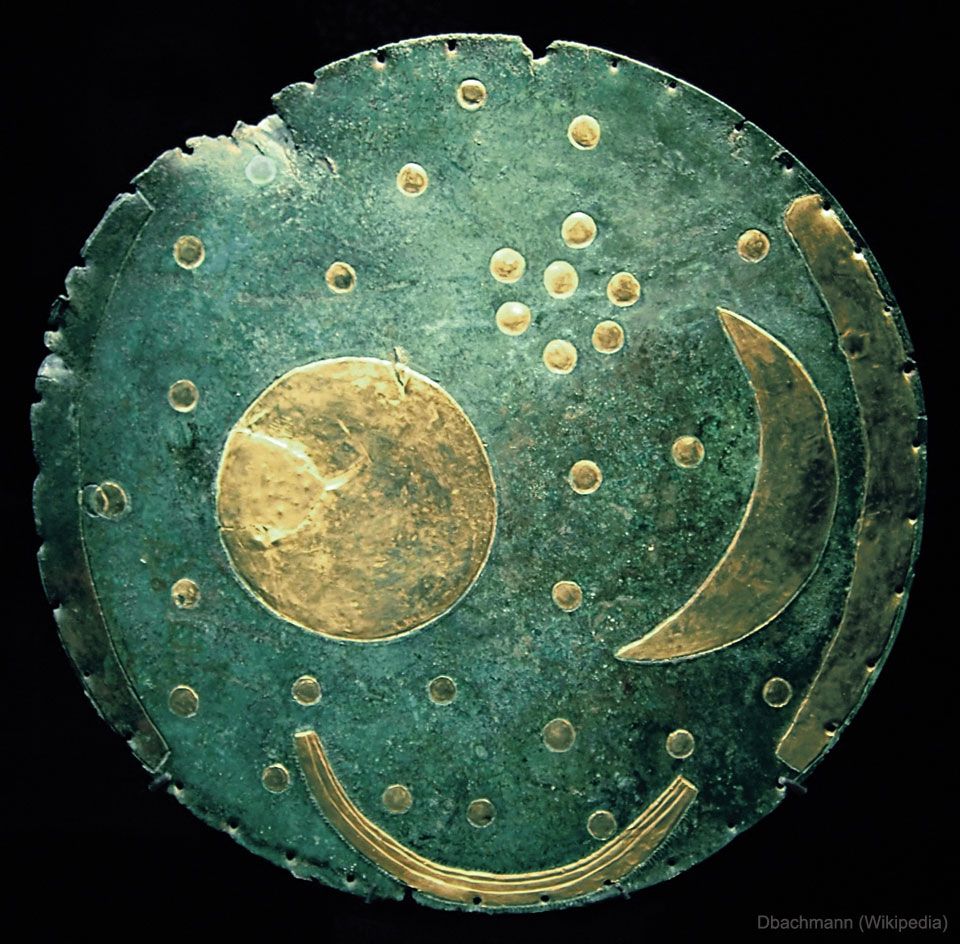 A circular bronze disk, about 30 cm across, with a dark green patina from age. Gold leaf inlays form a night sky: scattered round dots represent stars, a tight cluster of seven marks the Pleiades, a full circle near the center is the Sun, and a thin crescent below it is the Moon. Two gold arcs curve along opposite edges, possibly horizons or solstice markers; a third arc at the bottom may indicate a boat or river. The surface shows faint hammer marks and corrosion.