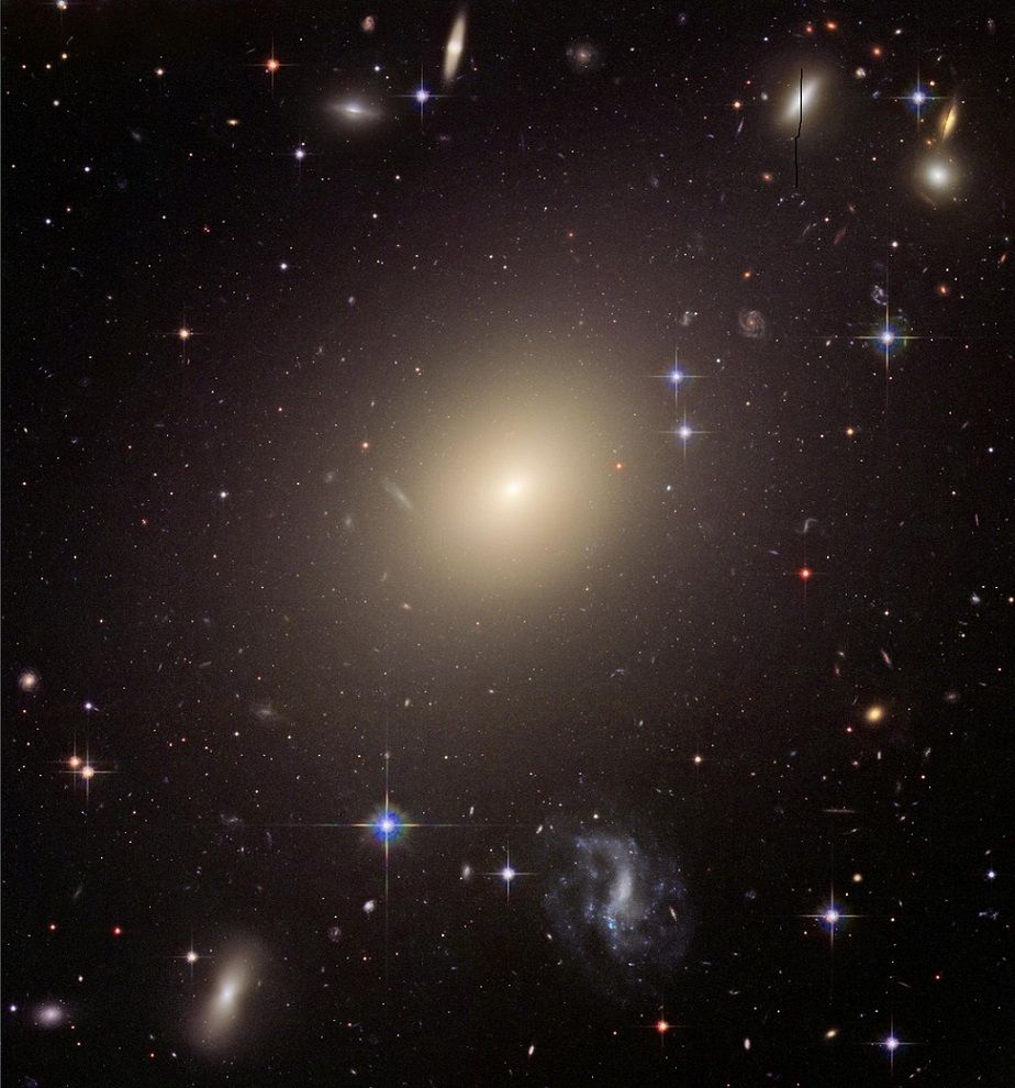 This Hubble image depicts the massive elliptical galaxy ESO 325-G004. Located about 450 million light-years from Earth in the Abell 3574 supercluster, it is an example of early elliptical galaxies (ETGs), which, billions of years ago, were like "cosmic fires" thanks to intense bursts of star formation. Their light, absorbed and re-emitted by cosmic dust, may have mixed with the CMB, the "roar" of the Big Bang, influencing what we observe today

During their formation, these galaxies shone 10,000 times brighter than they do today, contributing to the cosmic microwave background. Imagine a vast, luminous cloud suspended in a dark void, like a giant lantern illuminating the silence of space. The main galaxy has an oval shape, with a central core emitting a bright light, like the warm glow of a fire. This light gradually fades towards the edges, as if the galaxy were shrouded in a soft mist fading into darkness. The hue is warm, similar to a pale yellow or pale white, conveying a sense of  calm, like a celestial object that witnessed the birth of the universe. There are no sharp details or irregularities, but a smooth, uniform surface, conveying an impression of order and stability. Surrounding the galaxy, the background is dark, like a moonless night sky, dotted with specks of light, resembling distant sparks. Other galaxies are visible, scattered like cosmic dust in a vast empty space.The stars marked by bright rays (diffraction spikes) belong to our galaxy, the Milky Way, and are foreground stars that appear in the field of view of the image taken by Hubble. The image evokes a sense of grandeur and mystery, as if we were mentally touching a remote corner of the early universe.
