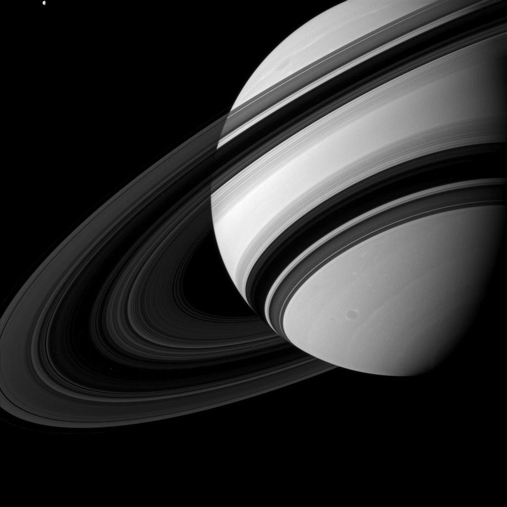 The image shows Saturn’s rings from their unlit side, where the light and dark areas are reversed compared to a typical view. The rings appear as concentric bands of light and dark gray, with the central B-ring looking darker than the brighter surrounding rings. The background is black space, with Saturn’s large, dark shadow on the right. At the top left, the moon Tethys is a faint, small bright dot. The image feels ethereal, with sharp contrasts between the rings and space.
