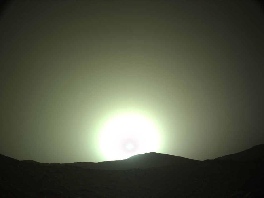 Martian sunset: the sun, low on the horizon, settles on the planet's dark, rocky profile like a huge, brilliant, whitish disk. Its outline, like the background, is greenish.