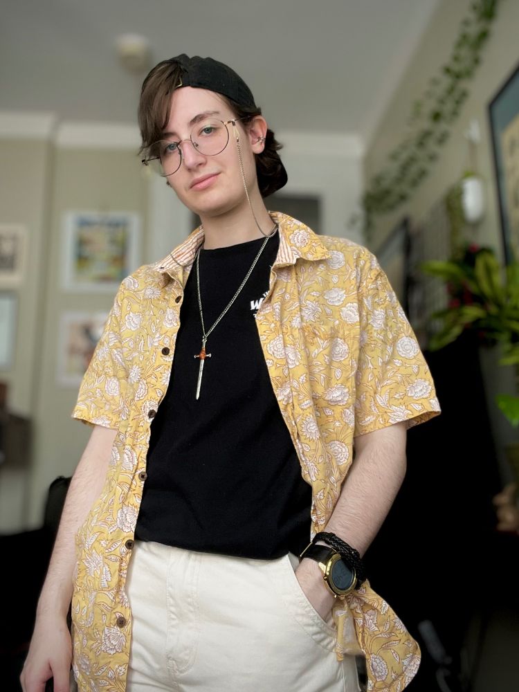 A photo of a person in a black tee, white pants, and with a yellow overshirt. They also have a black cap on backwards. They’re Caucasian with brown hair and blue eyes, and have one hand in their pocket while they look at the camera