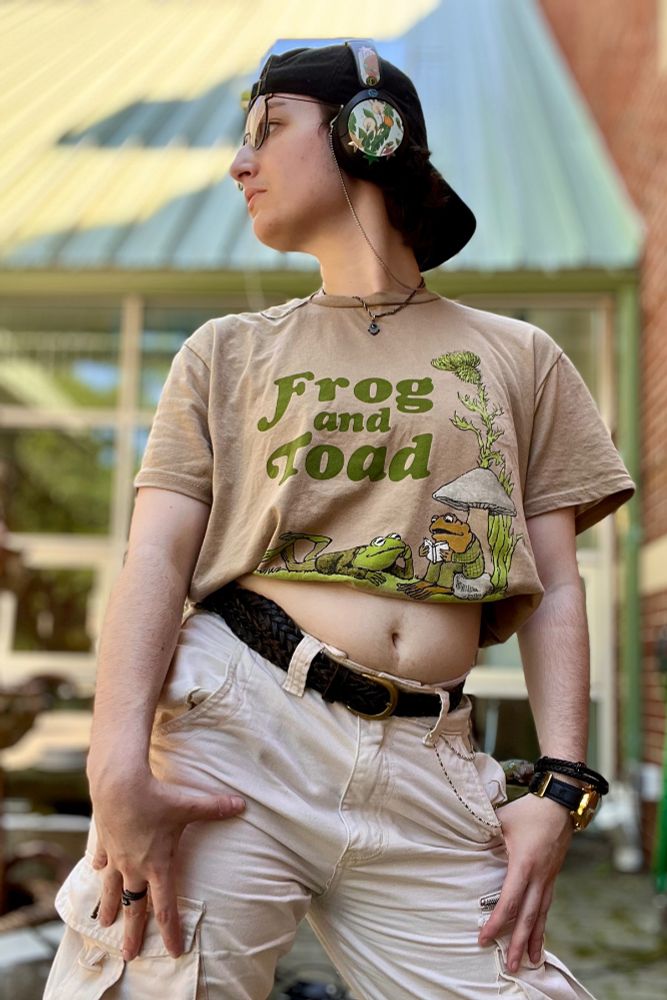 A masc nonbinary Caucasian person in cream colored cargo pants, a cropped brown Frog and Toad shirt, and a black snapback hat poses for the camera, looking off to the left. 