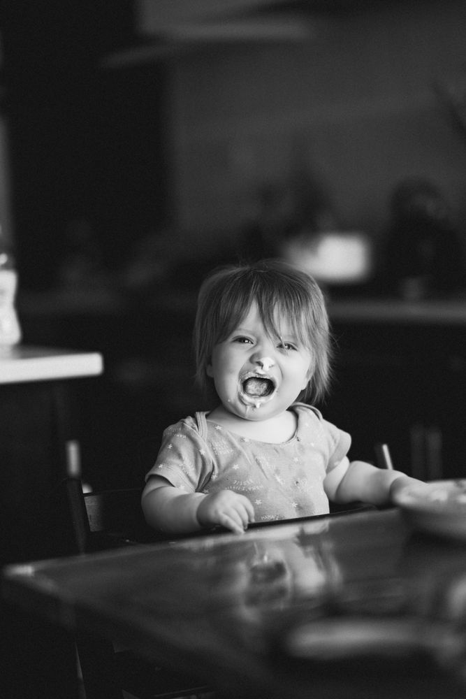 Bathed in the timeless elegance of black and white, this photograph captures a quiet, introspective moment of childhood. A young child sits at a table, their gaze gently directed toward something unseen, as if lost in thought. The soft, natural light plays across their features, accentuating the delicate contours of their face and the subtle texture of their short-sleeved, star-patterned shirt.

The background fades into a dreamy blur, an effect beautifully achieved with the Sony a7r5 and the exquisite 85mm f1.4 G Master lens. The shallow depth of field isolates the subject with precision, pulling the viewer into the quiet intimacy of the scene. The reflective surface of the table adds depth, mirroring hints of light and shadow, while scattered objects—indistinct but present—suggest the warmth of a lived-in space.

This image is a study in softness and nostalgia, evoking the simplicity and wonder of childhood. The monochrome tones strip away distractions, leaving only pure emotion and composition. It's a frame that speaks in hushed tones, inviting the viewer to pause and appreciate the beauty in the everyday. 
