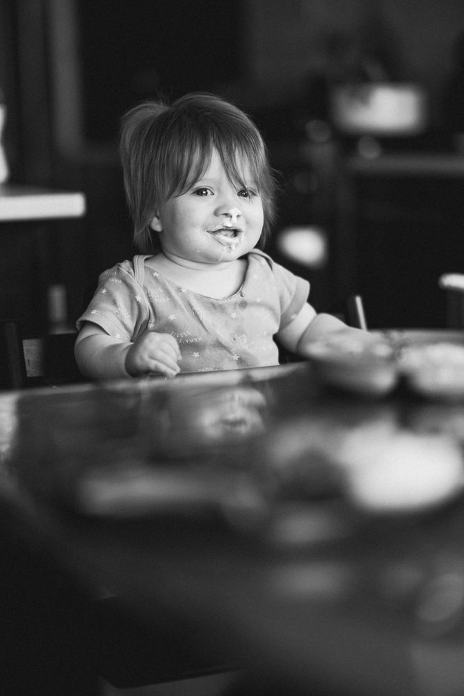A young child sits at a table, lost in thought, their gaze soft and introspective. Gentle natural light highlights their features, while the 85mm f1.4 G Master lens creates a dreamy blur in the background. The monochrome tones strip the scene to its essence—quiet emotion and simplicity.  