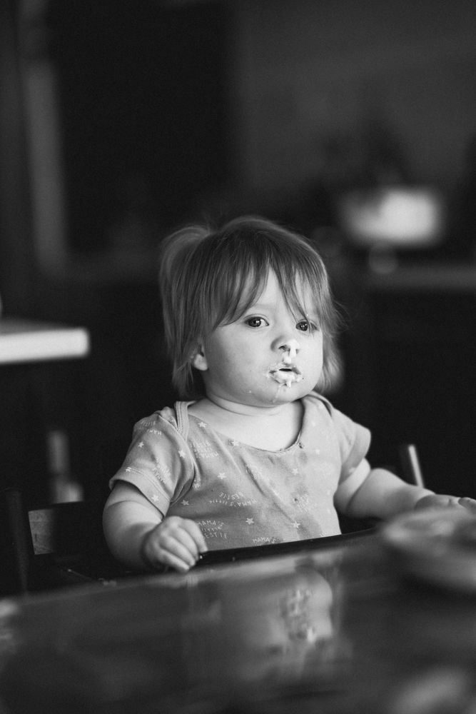 A young child sits at a table, lost in thought, their gaze soft and introspective. Gentle natural light highlights their features, while the 85mm f1.4 G Master lens creates a dreamy blur in the background. The monochrome tones strip the scene to its essence—quiet emotion and simplicity.  