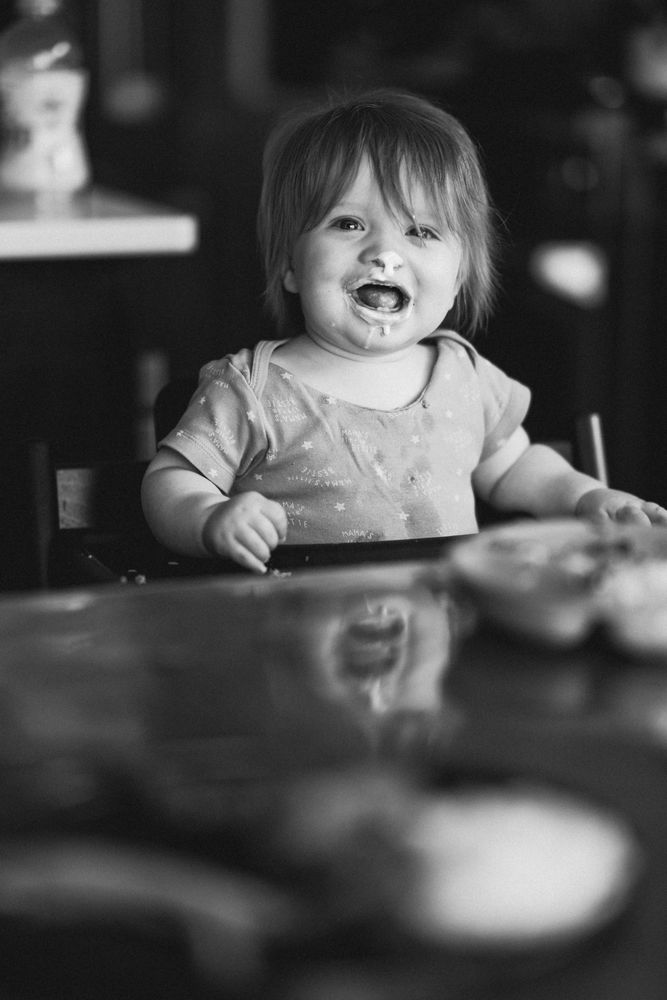 A young child sits at a table, lost in thought, their gaze soft and introspective. Gentle natural light highlights their features, while the 85mm f1.4 G Master lens creates a dreamy blur in the background. The monochrome tones strip the scene to its essence—quiet emotion and simplicity.  