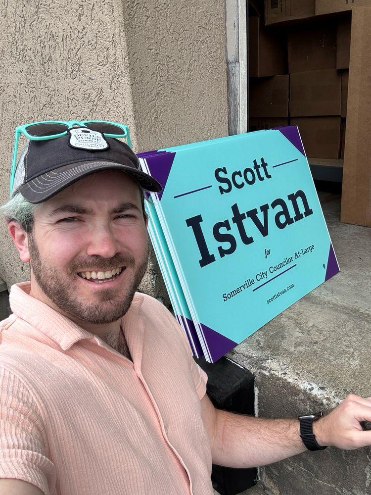 A selfie of me (mid 30s bearded white guy with teal hair wearing a baseball hat) next to a teal and purple campaign yard sign that reads “Scott Istvan for Somerville city councilor at large”