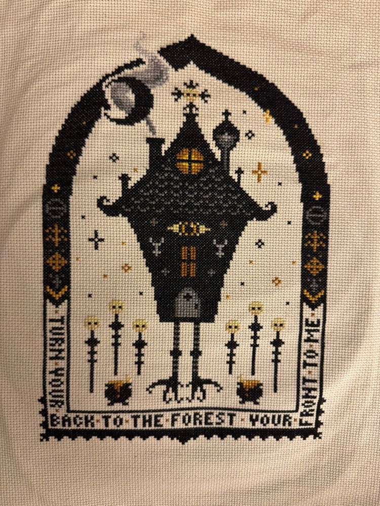A cross stitch of a hut on chicken legs with an eye for a window. There are skulls on posts and the words “Turn your back to the forest, your front to me”