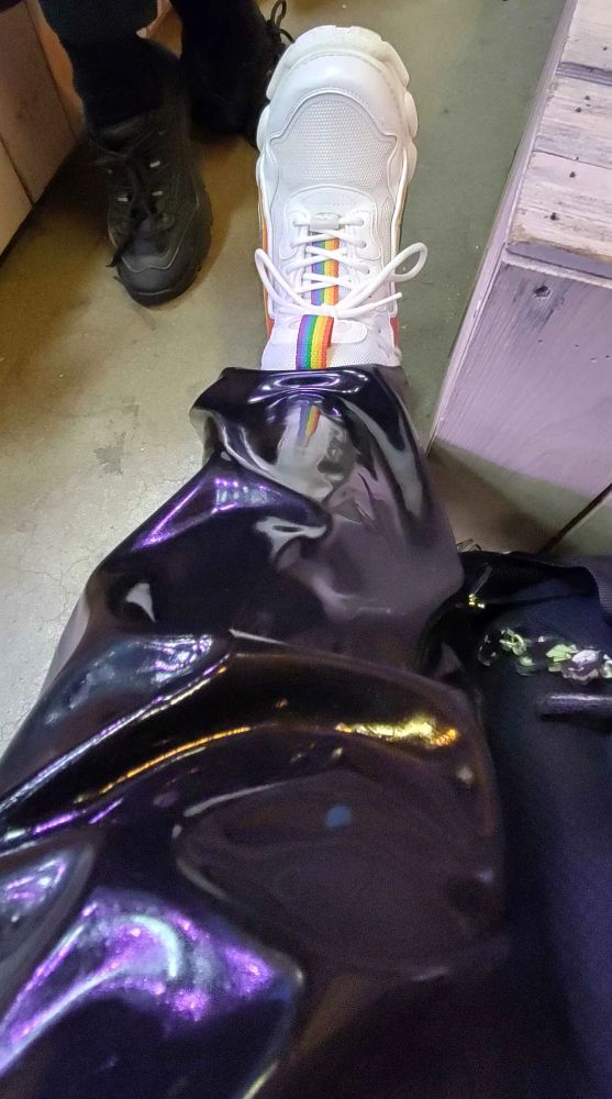 Photo down my spread leg, showing one of my white Buffalo boots with rainbow-flag stripes on the tongue and the sides of the boots. My leg is covered in latex by my loose fitting latex pants. Multiple purple, light blue, yellow and orange reflections from the ceiling lights are visible. Down the lower leg a mirror reflection of my boot is visible, including the rainbow strip.