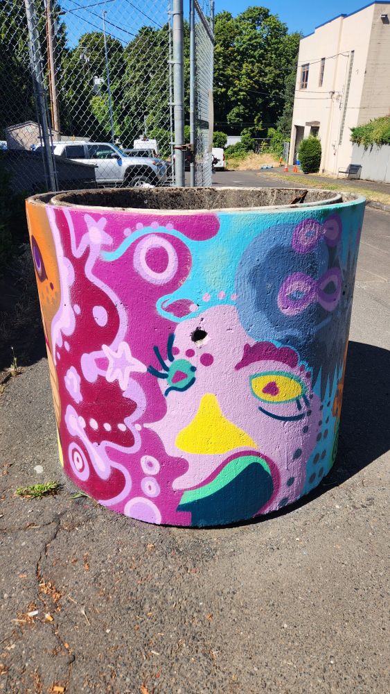 A concrete barrel planter with an abstract face in pink, yellow and blue. 