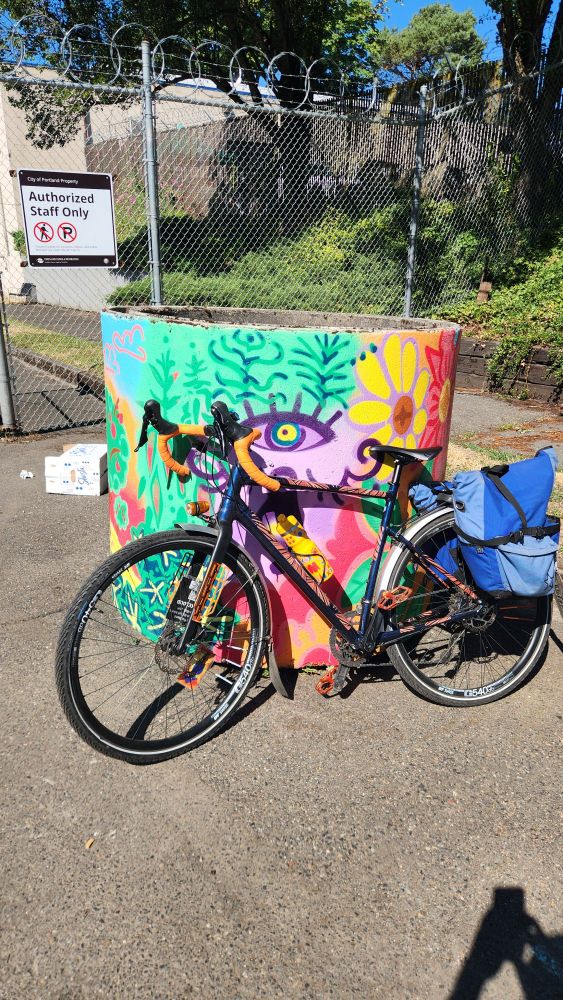 A concrete barrel planter painted with colorful flowers and trees. A bike is leaning on the barrel. 