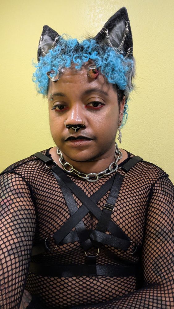 Selfie of Silas IRL. He is wearing a long sleeved fishnet top with a black, inverted pentagram harness over the shirt and a thick metal chain around his neck. He has bright blue hair and small floral horns on his forehead. He is wearing black, pierced wolf ears on top of his head. His upper lip is painted black as well as the underside of his nose.