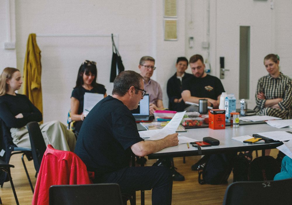 Photo of Mark Thomas in profile, sitting at a table reading aloud from a script. Members of the production team are visible in the background, sitting on chairs and listening in.