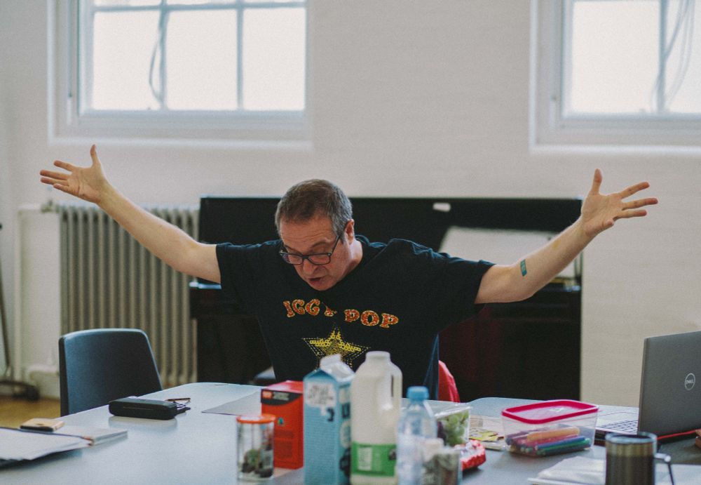 Photo of Mark Thomas sitting at a table in a rehearsal room, reading aloud from a script on the table in front of him. He's holding both arms up in the air in a gesture of emphasis.