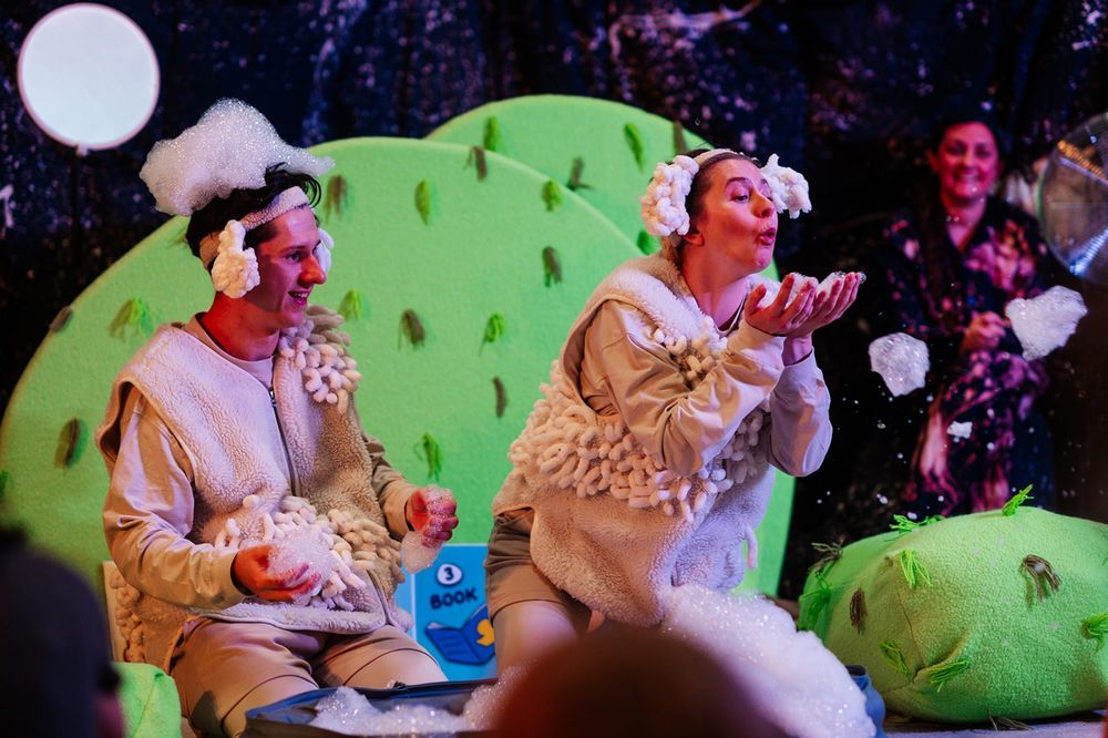Photo from a performance of 'Counting Sheeps', showing two performers dressed as sheep kneeling on stage with large green pieces of set resembling trees and grass behind them. They are blowing pieces of bubble foam from their hands into the air in a whimsical manner.