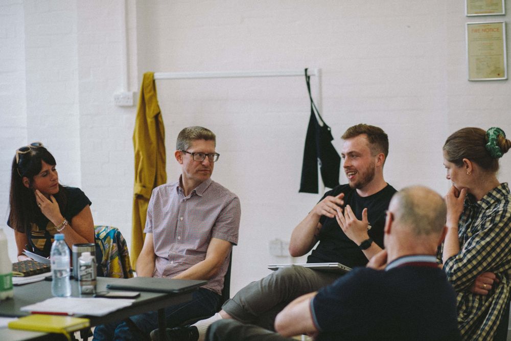 Photo of members of the production team sitting around a table in the rehearsal room, having a discussion.