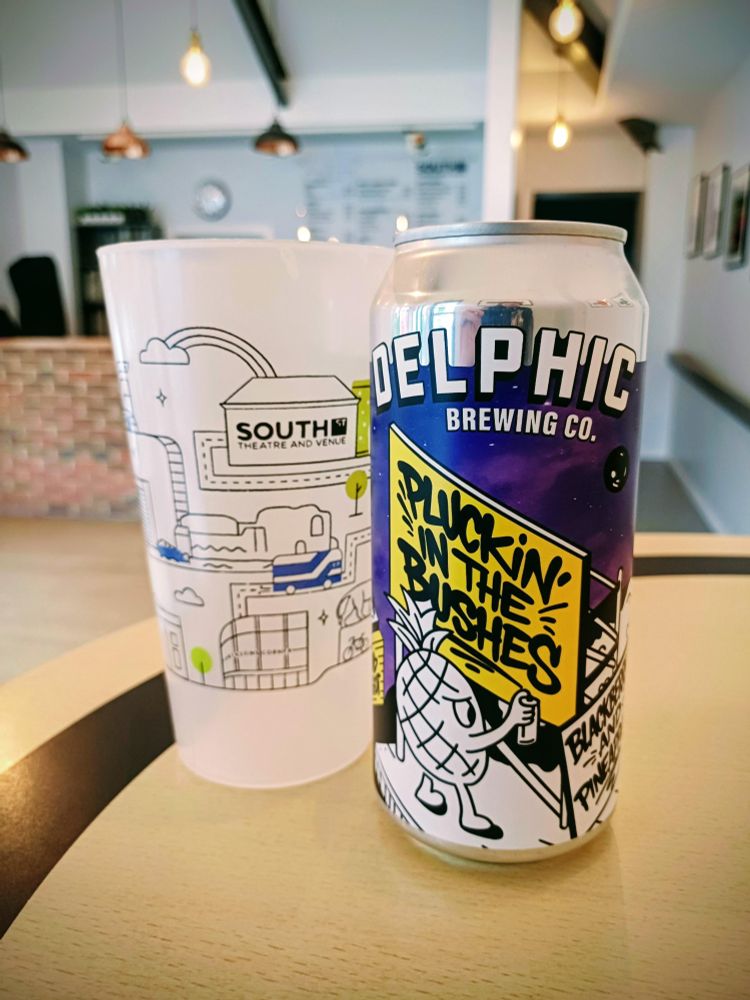 Photo of a can of Delphic Brewing Co's Pluckin in the Bushes beer, next to a South Street branded reusable cup, on one of the South Street Bar's tabletops. The South Street Bar room is visible and slightly blurry in the background.