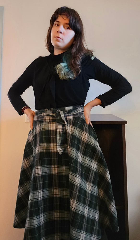 Jax looks into the camera. She wears a plaid skirt, a black turtleneck, and an overshirt with a pussy bow 