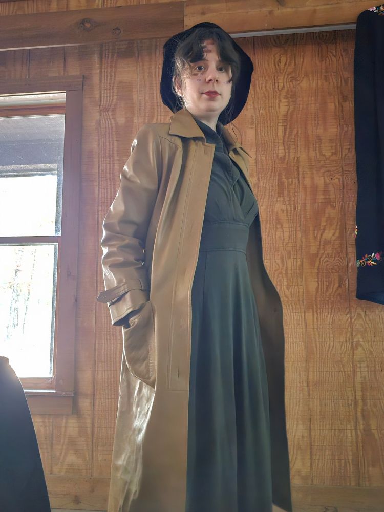 Jax in a long green dress and long coat and a brickwork scar on her face