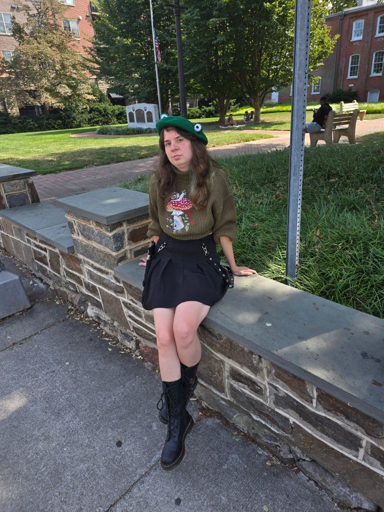 Jax sits on a low ornamental wall in a mini skirt and green frog beret and cropped sweater and docs