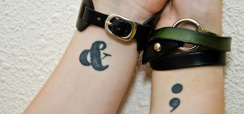 Close-up photo of two pale-skinned wrists against a white wall; the right wrist has a black semicolon tattoo and the left wrist has a black ampersand tattoo. Both wrists are wearing different leather cuffs with metal buckles.