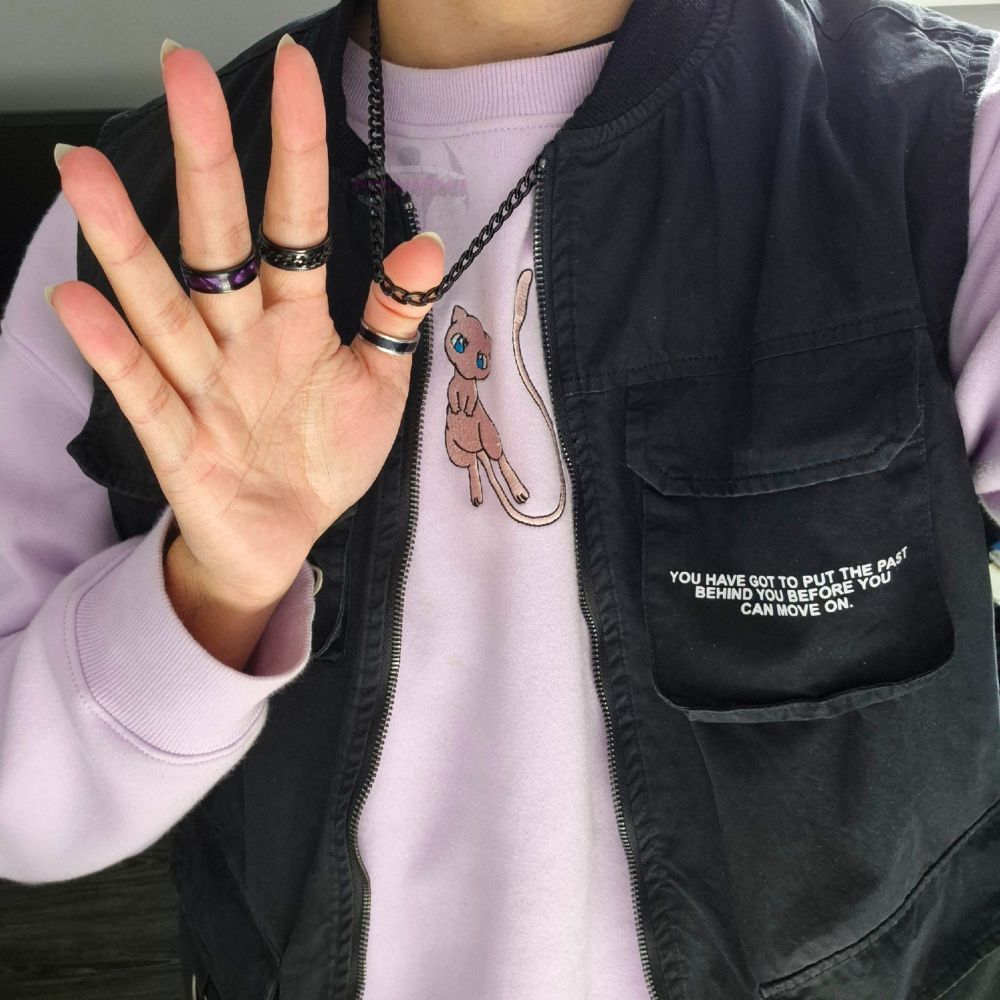 Pink Mew sweater from Pokemon. Black open cargotop  over it. Holding black necklace chain with thumb of hand where palm faces camera. Purple ring on indexfinger, black chain ring on indexfinger, black and silver ring in thumb.
