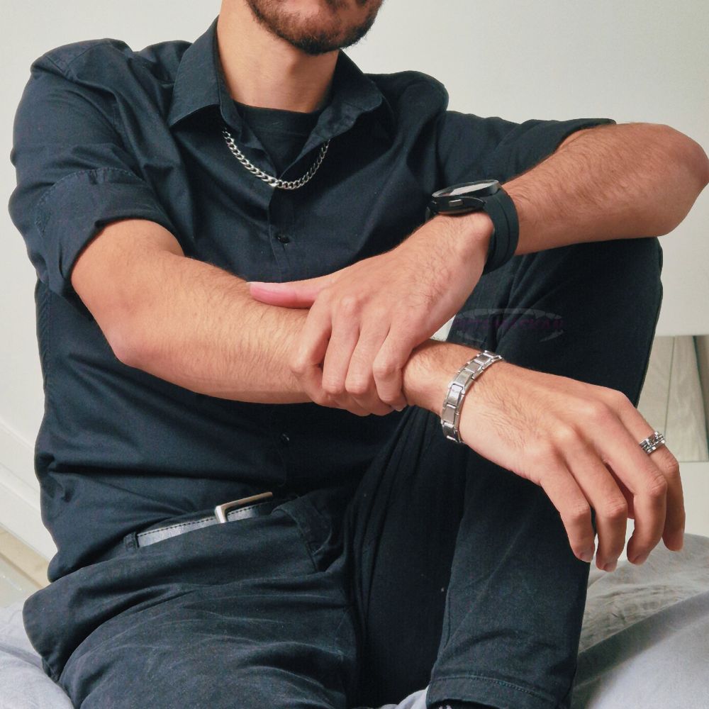 Black outfit, button up, silver chain, bracelet and ring, one knee up, holding forearm. 