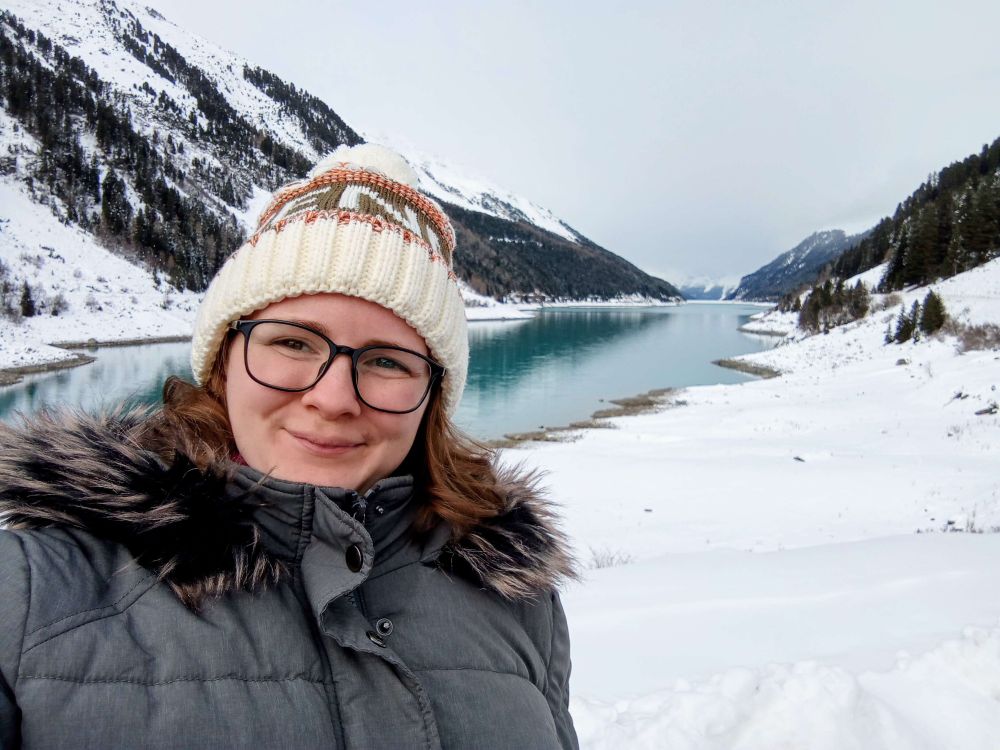 Photo of me with a white and green winter hat  and a grey winter jacket, wearing glasses and smiling in the camera. In the background is a landscape of snow, snowie mountains and a blue lake.