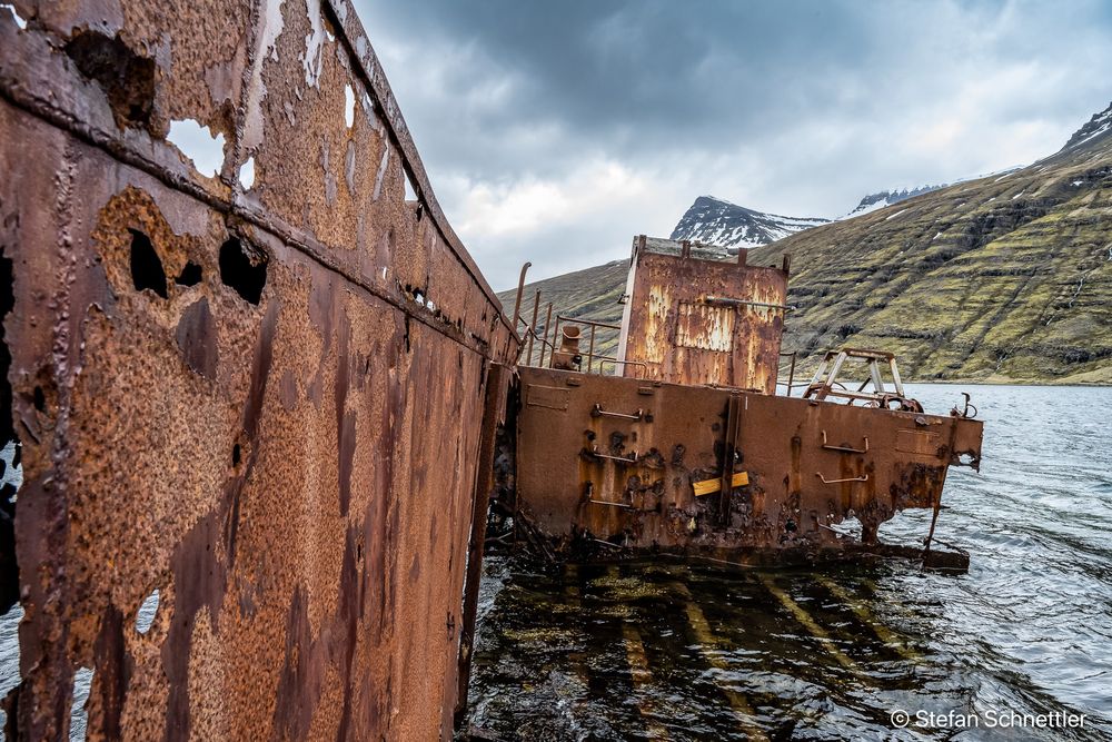 Rusting, hole-riddled shipwreck grounded in shallow water beside a fjord, with snow-dusted mountains under a brooding sky.