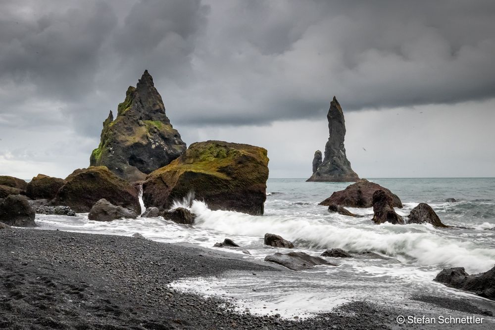 Dramatic coastal scene at Reynisfjara beach in Iceland, featuring jagged sea stacks rising sharply from the North Atlantic under heavy, brooding clouds. Dark volcanic basalt stacks, covered in patches of moss, scatter along the black sand beach in the foreground. Waves crash against the shoreline, their white foam contrasting starkly with the dark tones of sand and stone. The mood is raw and powerful, evoking the untamed spirit of Iceland’s southern coast.