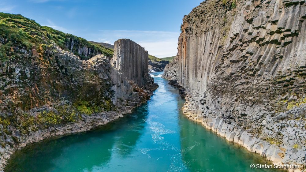 Wide view of Stuðlagil Canyon, East Iceland. The turquoise Jökla River runs between towering hexagonal basalt columns on both sides; broken blocks and patches of green moss line the base. Bright summer light under a clear blue sky, no people.