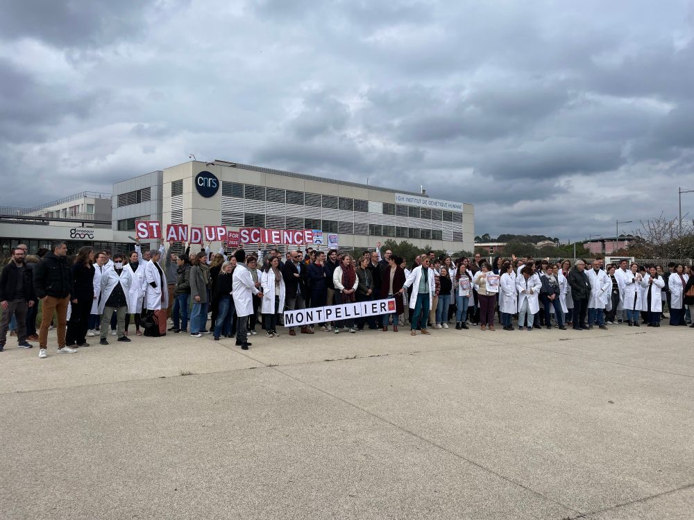 Photo d'un regroupement de plusieurs dizaines de scientifiques avec et sans blouses, en plan large, posant devant le campus Arnaud de Villeneuve à Montpellier-Occitanie, avec des pancartes Stand Up for Science.