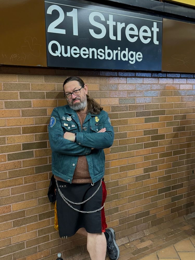 Me leaning against the wall, below the “21 Street Queensbridge” sign in said subway station. Wearing my Gothic pride, kilt, and my most recent decorated denim jacket creation.