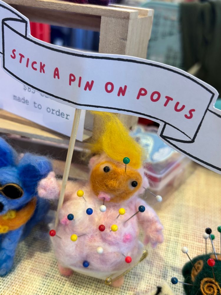 A needlefelted baby trump voodoo doll full of pins holding a sign that says “stick a pin on potus” craftersofbluesky activism 