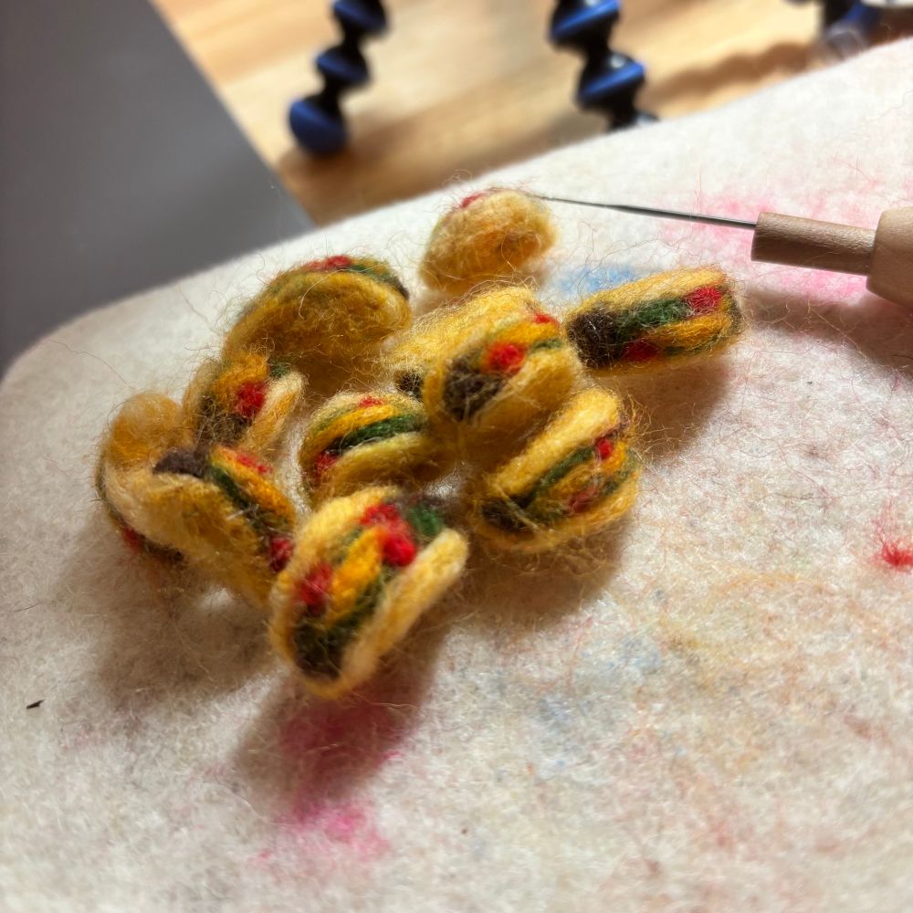 A pile of little teeny tacos hand crafted from wool on a needlefelting mat with a tool in the background🧶 