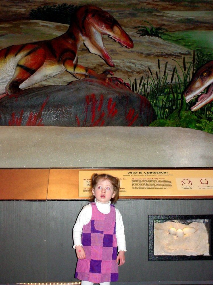 me as an adorable child, in a cute pink and purple outfit with a white turtleneck, standing beneath a dinosaur animatronic exhibit at the pacific science center 