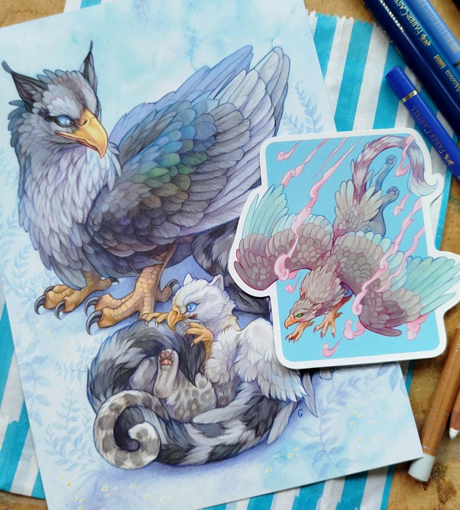 Photo of a griffon parent and chick art print and a swooping griffon sticker surrounded by pencils. 