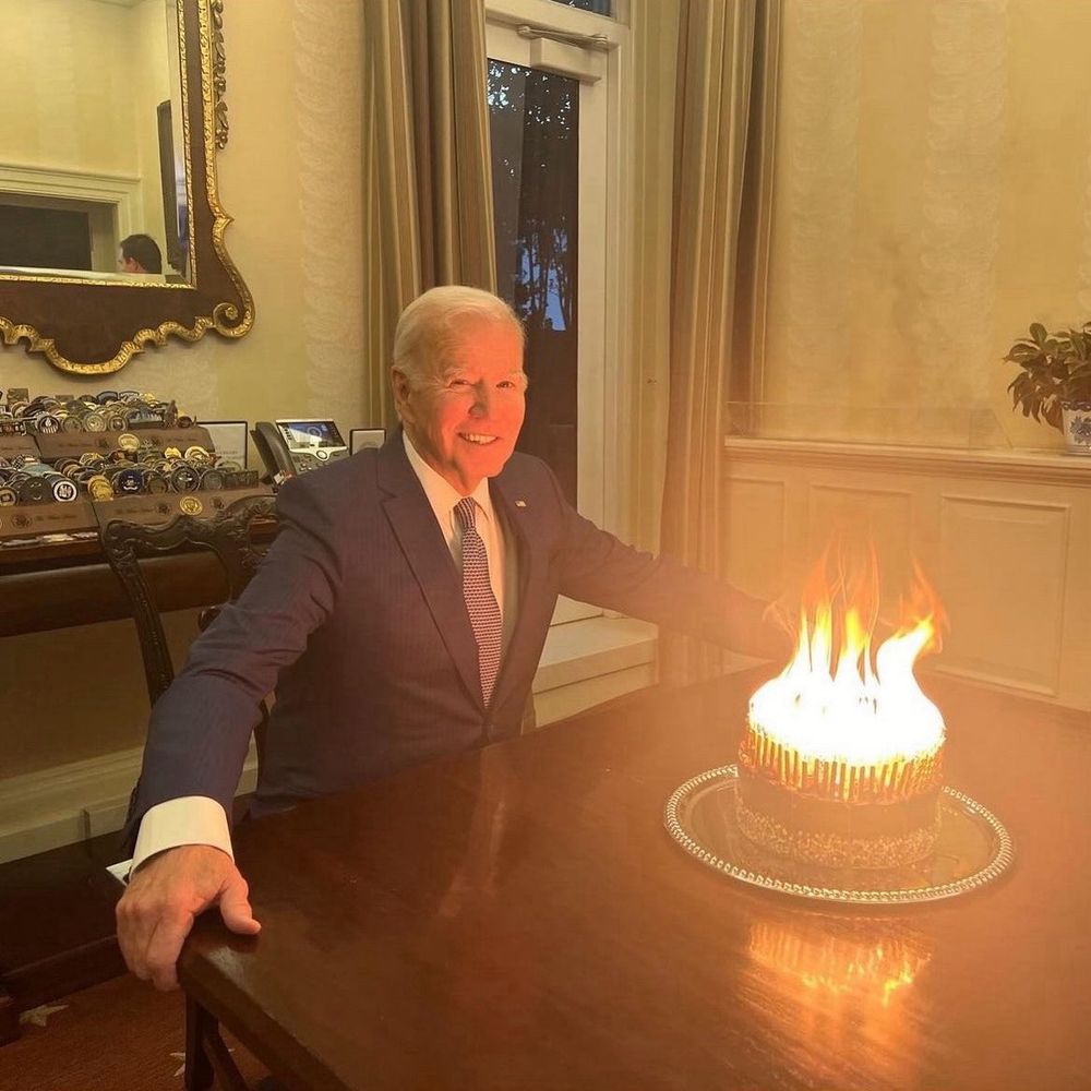 Joe Biden sits at a big table, smiling at the camera with a flaming birthday cake in front of him.