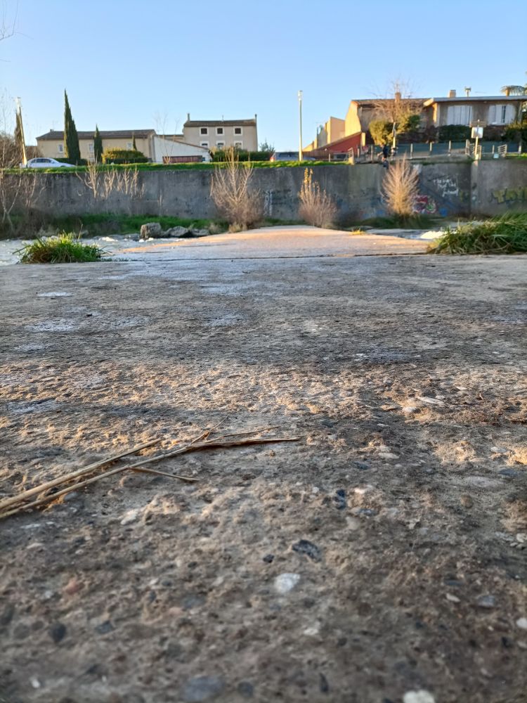 Un sol à la texture caillouteuse semble aller vers un mur en total perpendiculaire. Il y a de l'eau au bout, à droite et à gauche du chemin. La ville est derrière le mur perpendiculaire et la lumière est rasante.