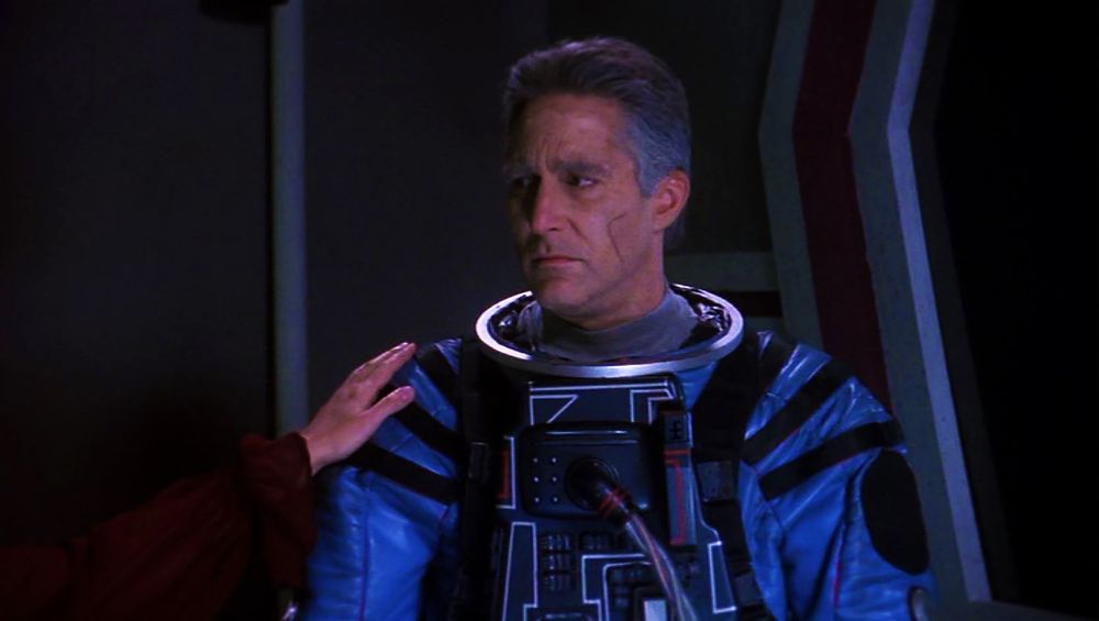 A suddenly aged and scarred Jeffrey Sinclair in a blue spacesuit looks regretful, comforted by a hand on his shoulder
