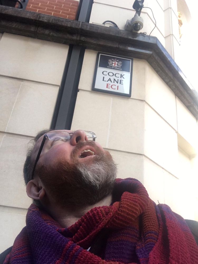 Me in glasses, beard and scarf, looking up at a street sign for COCK LANE EC1