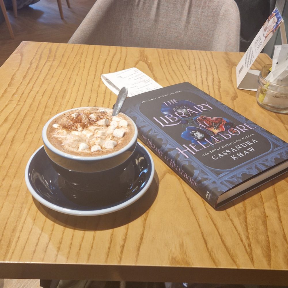 A mug of hot chocolate with marshmallows on a tablext next to a book. The book is The Library at Hellebore by Cassandra Khaw.