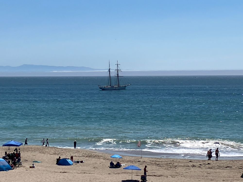 A two-masted brigantine (a “tall ship”, though on the smaller side) lies at anchor offshore from a sandy beach. A shallow fog bank lies well offshore, with the top of some hills poking out above. Some people may be seen on the beach, with a variety of umbrellas and other shade structures.