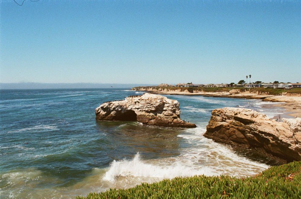 The natural bridge with a gap between it and a rock outcrop (there used to be a second natural bridge, it fell down years ago). Waves may be seen breaking at the bottom of the photo, along with invasive ice plant. A point in the background has buildings and various vegetation on it, including two tall palm trees. 28mm setting on the lens.