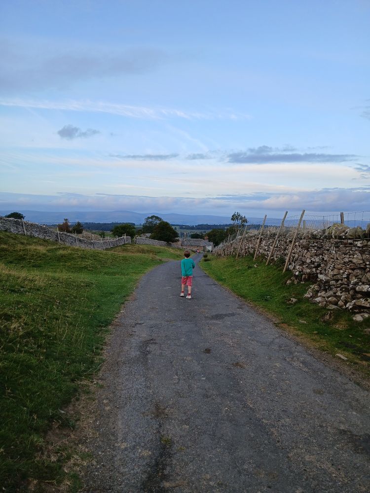 A small boy runs down a fell top road, edged with a drystone wall, with a view of distant fells.