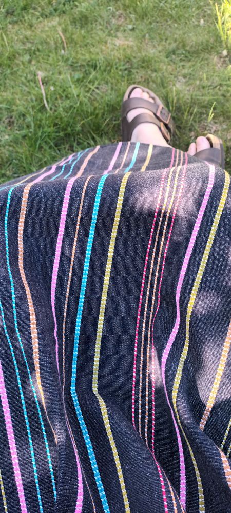 A pair of white feet wearing brown sandals emerging from a navy skirt with thin stripes of bright blue and pink, yellow, brown, and red.
