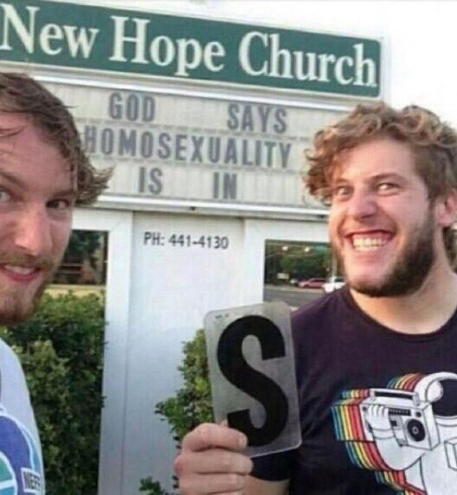 A sign in the background says New Hope church, God says homosexuality is in. In the foreground there are two men smiling holding the letter s that they've taken from the sign