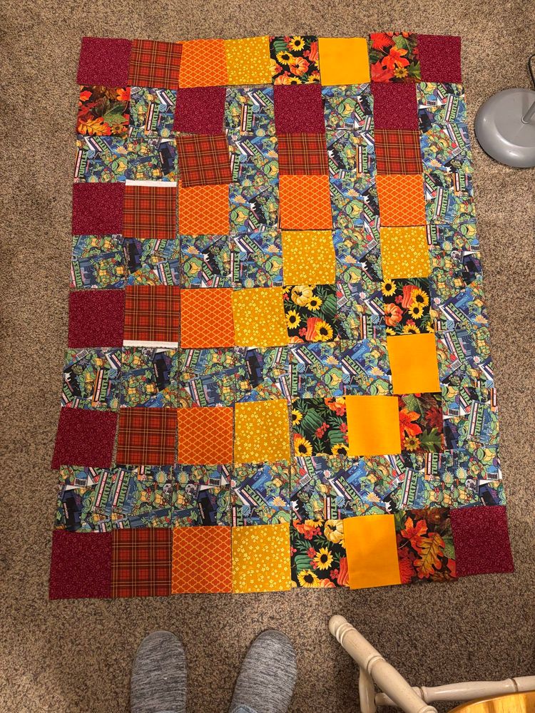 Quilt pattern I have laying out on carpet. Pattern is using teenage mutant ninja turtle print with warm / autumn colors: floral maroon, orange / red plaid, orange with a yellow pattern on it, gold / yellow flowers, black print w/ sunflowers & pumpkins, golden orange, and autumn leaves print squares. 