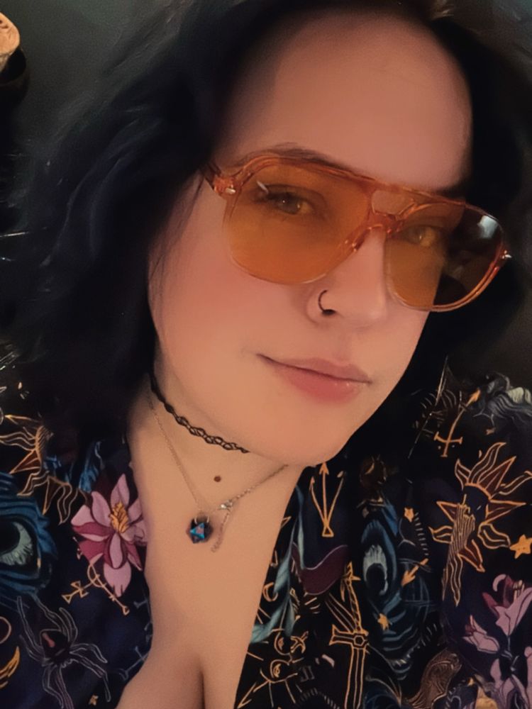 Selfie of the user wearing orange-colored sunglasses in a similar style to the ones seen in the game, Date Everything. They also are wearing the Critical Role Mollymauk patterned button-down shirt & a d20 necklace.