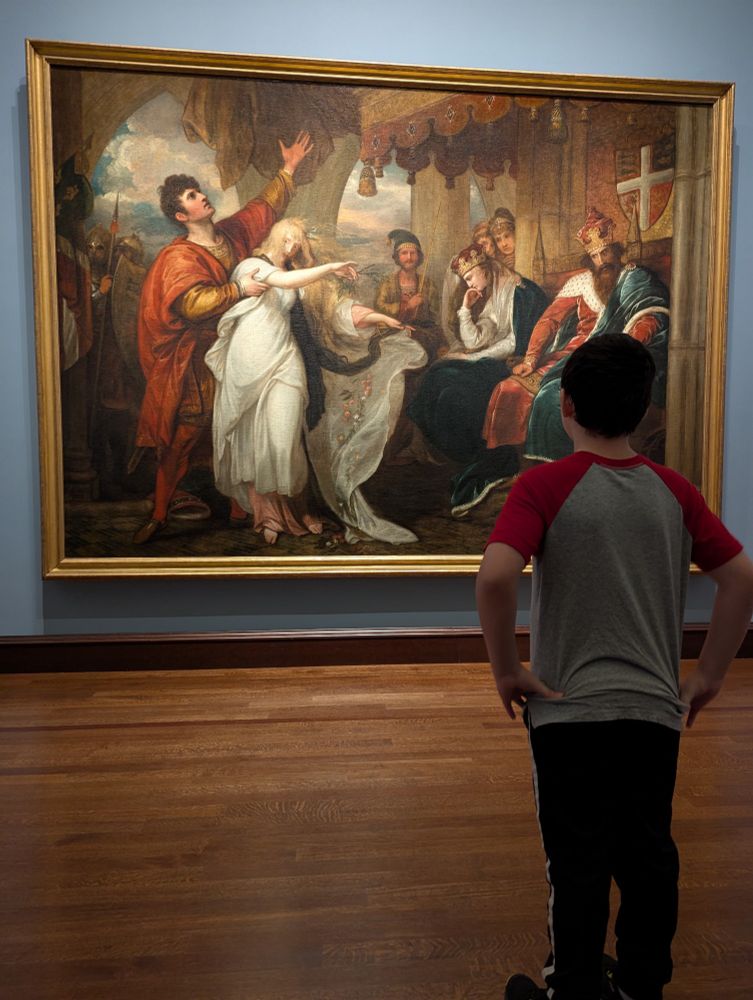 A young male child stands in front of a large oil painting depicting a scene from Hamlet.